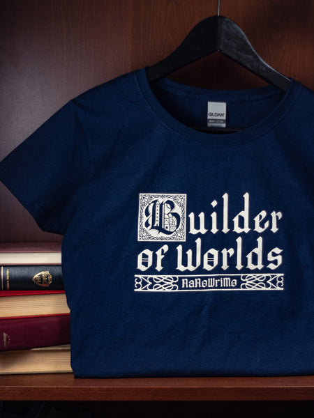The 2022 Camp shirt is navy with a simple, cream white print. The design features the text "Builder" on one line, and "of Worlds" on a second line, written in a Blackletter style font. The "B" is illustrated with more detail as though it were a drop cap in an illuminated manuscript. Below the second line, "NaNoWriMo" is centered and surrounded by more line-design flourishes.