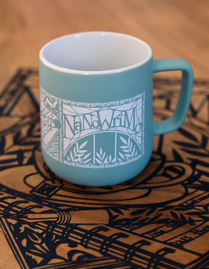Teal mug with white papercut-style print of a cup of tea, fountain pen, and stack of books. The NaNoWriMo name sits on top of the image. Botanical leaves and filigree fill the background. The image wraps around the mug in three panels. 