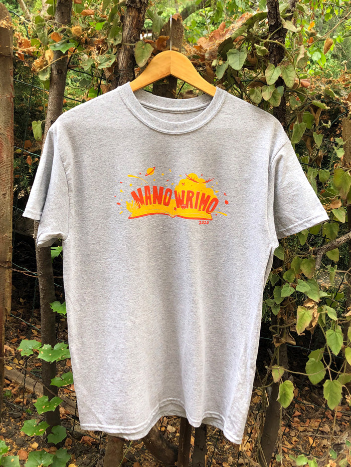A heather gray T-shirt features an open book with a red cover and yellow pages. The words “NaNo” and “WriMo” pop out of the book in red, with “2020” in red under the book. Red rose buds, a yellow and red deer, a yellow cloud, a small planet, and paint splatter all burst out from the book.