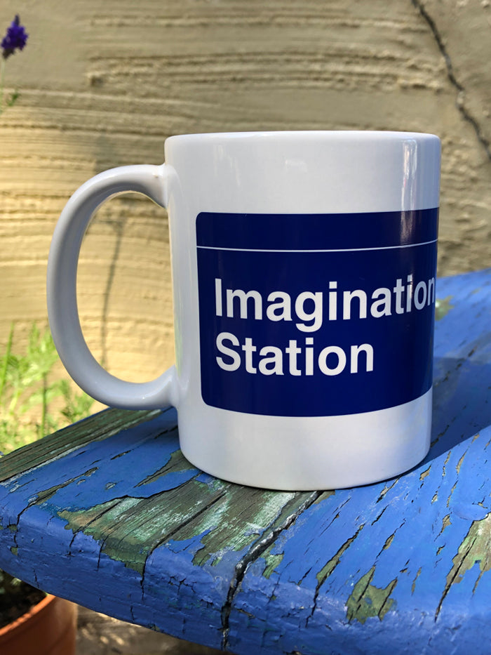 A white mug with a dark blue wrap-around print. One side of the mug says "Imagination Station" in a white font that evokes classic subway station signage. The other side has four lighter blue circles with the letters N, A, N, O in their center in white and in the style of NYC subway line signage, with the word "NaNoWriMo" in smaller, white print below the circles. There is a thin white line going across the top of the dark blue print, above the text on both sides.