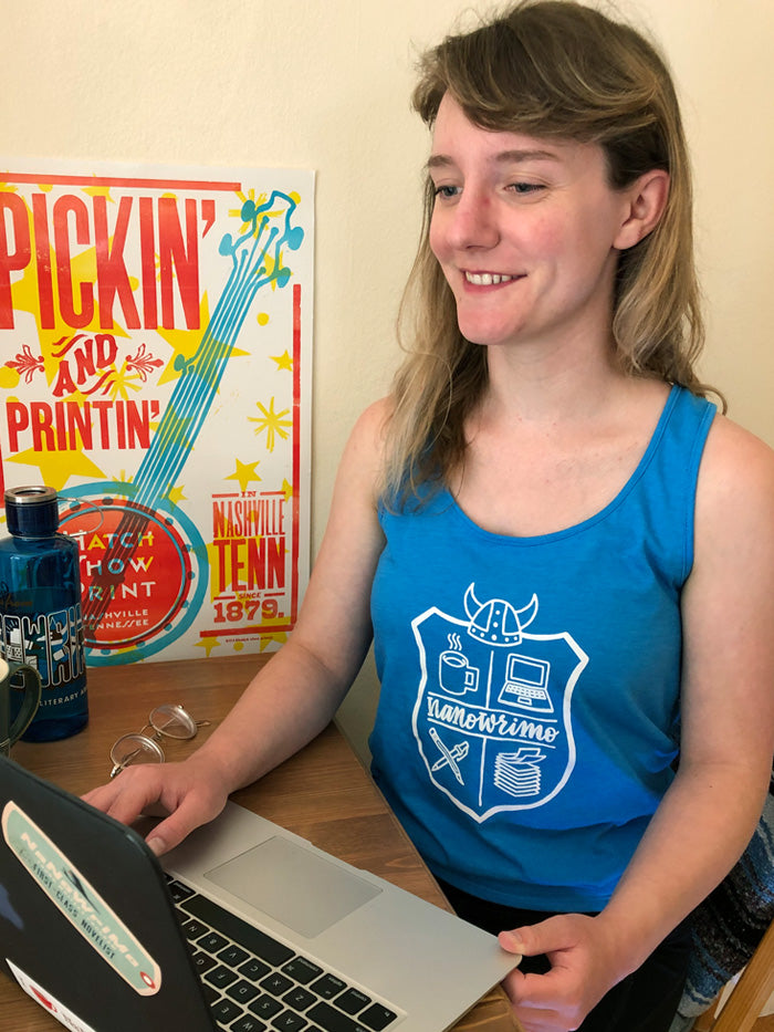 This tank top is turquoise blue with a white print. The design features a stylized NaNoWriMo crest logo, which includes a Viking helmet on top of shield with a coffee cup and laptop computer in the top quadrants and pens crossed and a stack of papers in the bottom quadrants. The text "NaNoWriMo" separates the top and bottom in a cursive typeface.