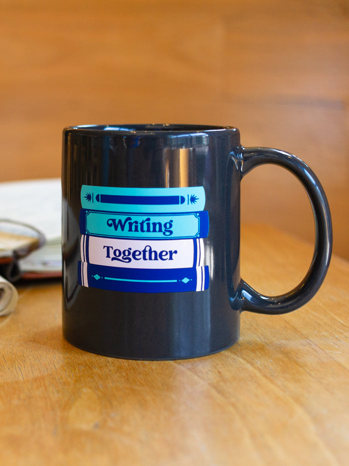 This charcoal gray mug is printed with teal, dark blue, and pink ink. One side includes a stack of four books with the spines facing out, and the center two books have "Writing" and "Together" printed on the spines. THe other side features a closed planner with "NaNoWriMo" written across the center, and a pen and pencil sitting next to it slightly askew.